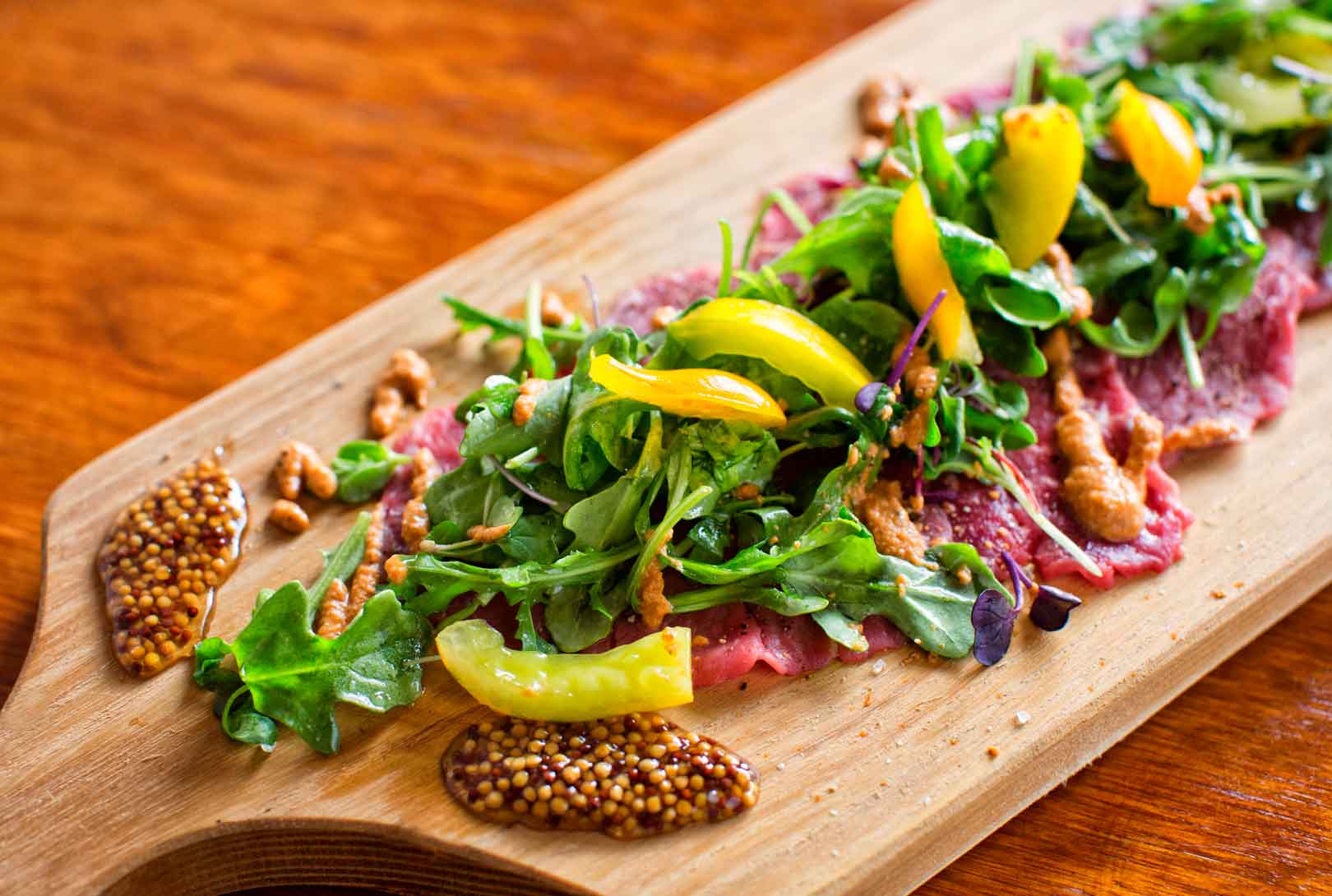 A cutting board with beef carpaccio topped with arugula peppers and jam served as appetizer