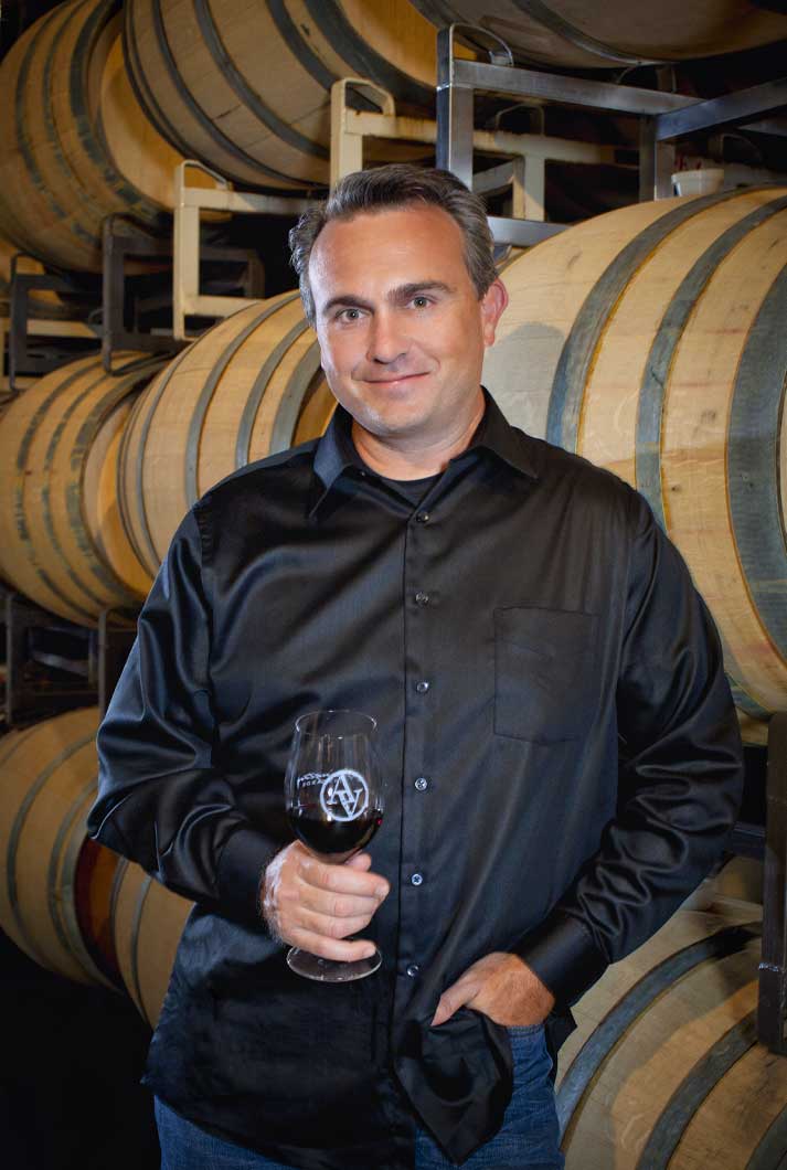 Kip Summers wearing black dress shirt holding glass of red wine standing near wine barrels at winery