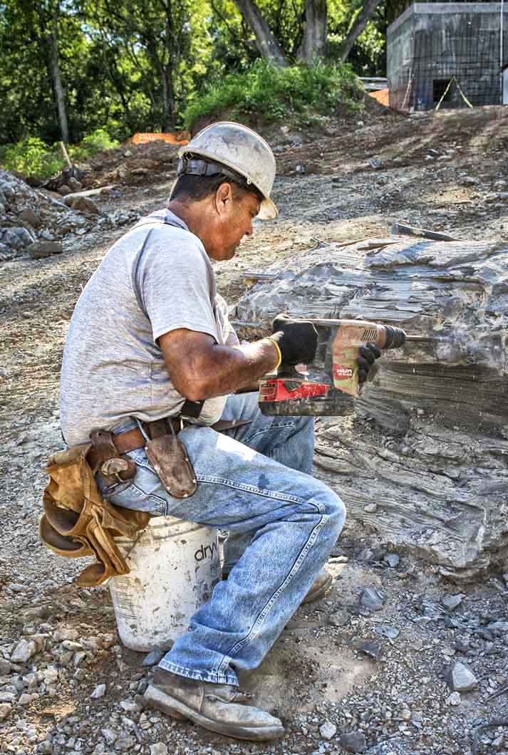 a man sitting on white bucket drilling into concrete fabricated rock