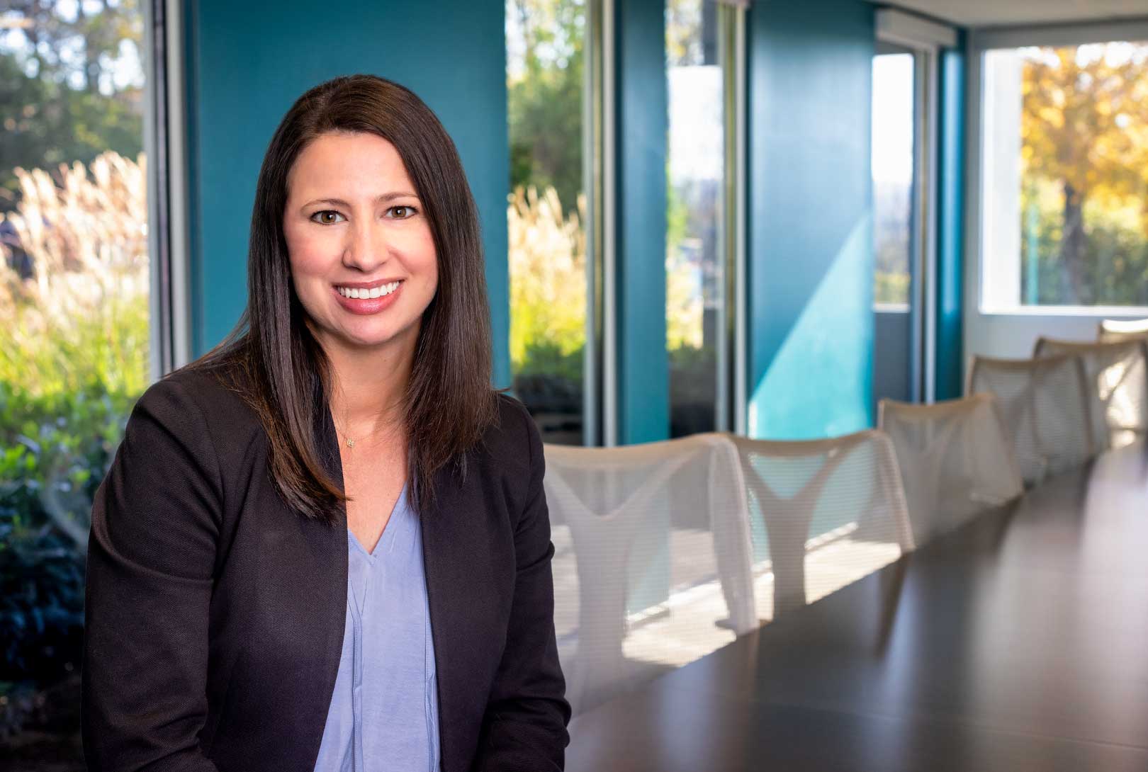 executive headshot of Meg Chamblee in a conference room leaning on a table with white chairs and blue walls