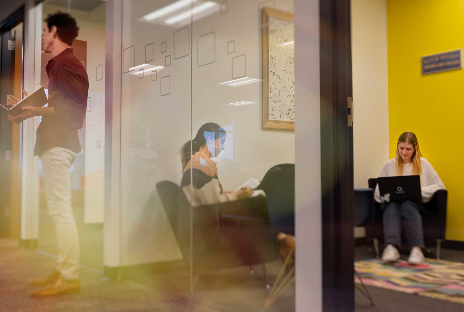 two female employees working on laptops in office space, others in hall through glass