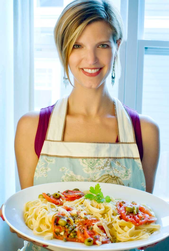 health coach Amber Robertson in apron holding a pasta dish with tomatoes and olives