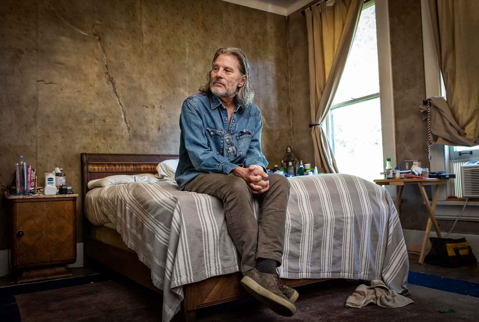 Buddy Jackson wearing a blue jean shirt, corduroy pants, and sneakers sitting on his bed