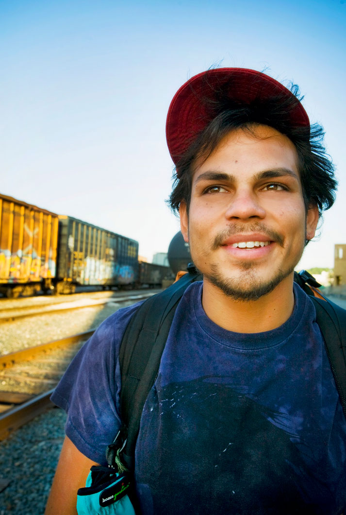 a young male hobo train jumper in a red ball cap and blue shirt on railroad tracks near freight cars