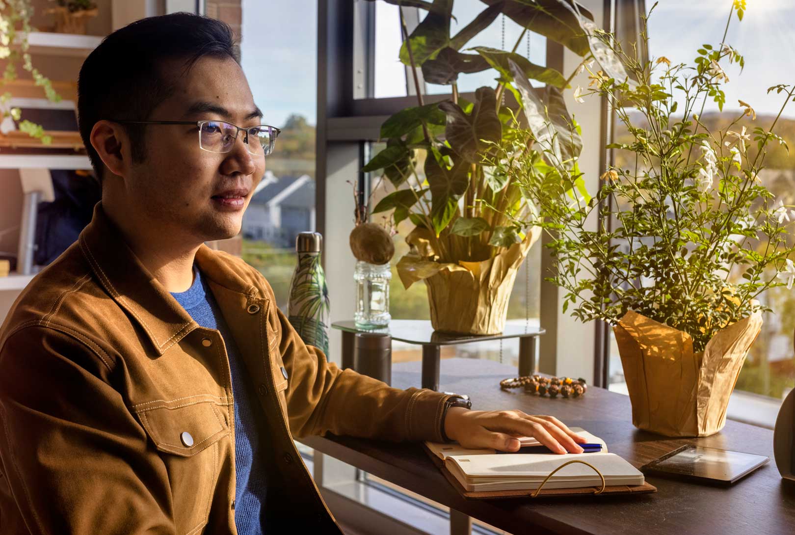 Li Qin in his home sitting near a window with plants and a journal