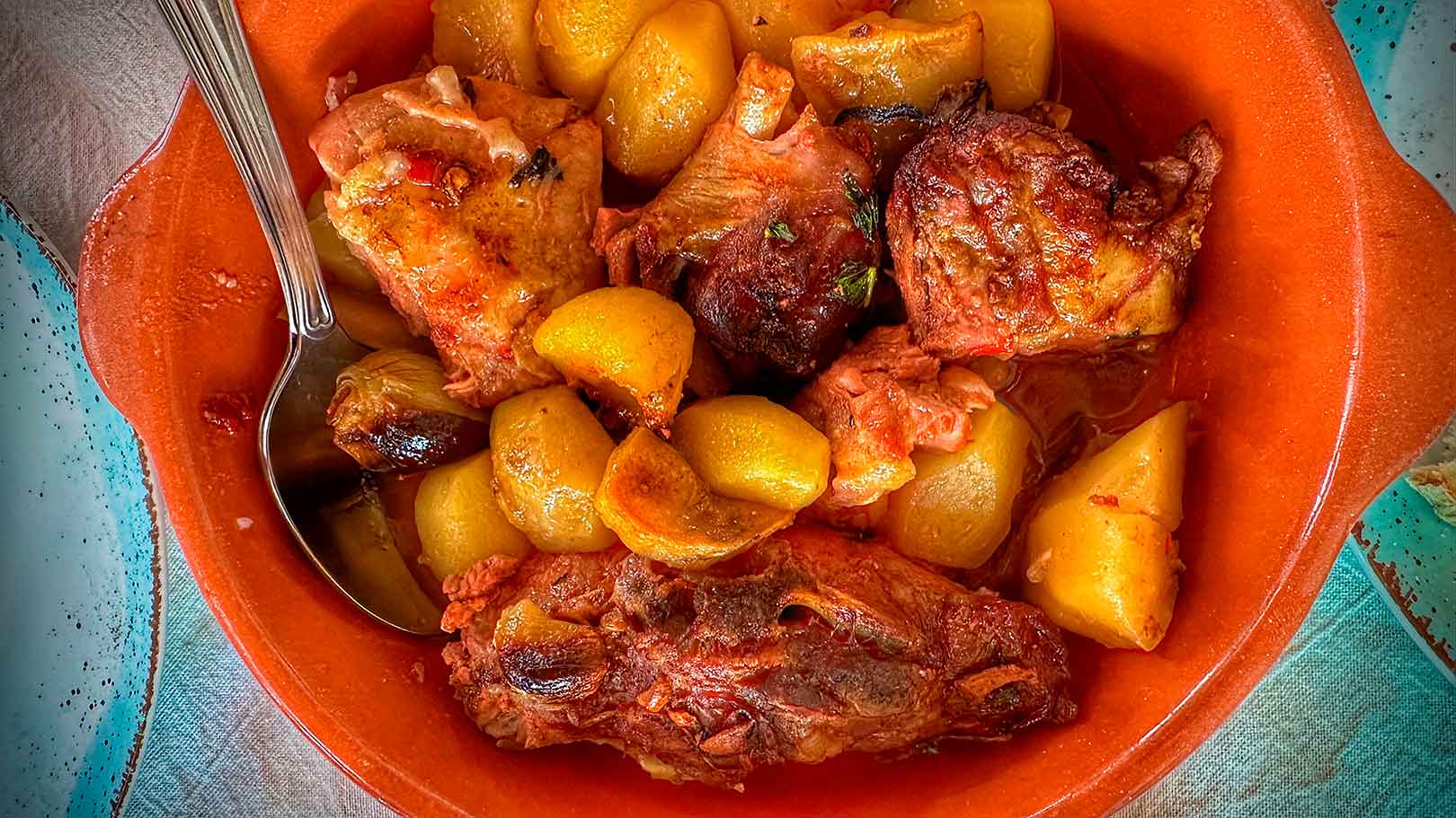 A clay bowl filled with roasted lamb meat and potatoes, accompanied by a metal spoon.