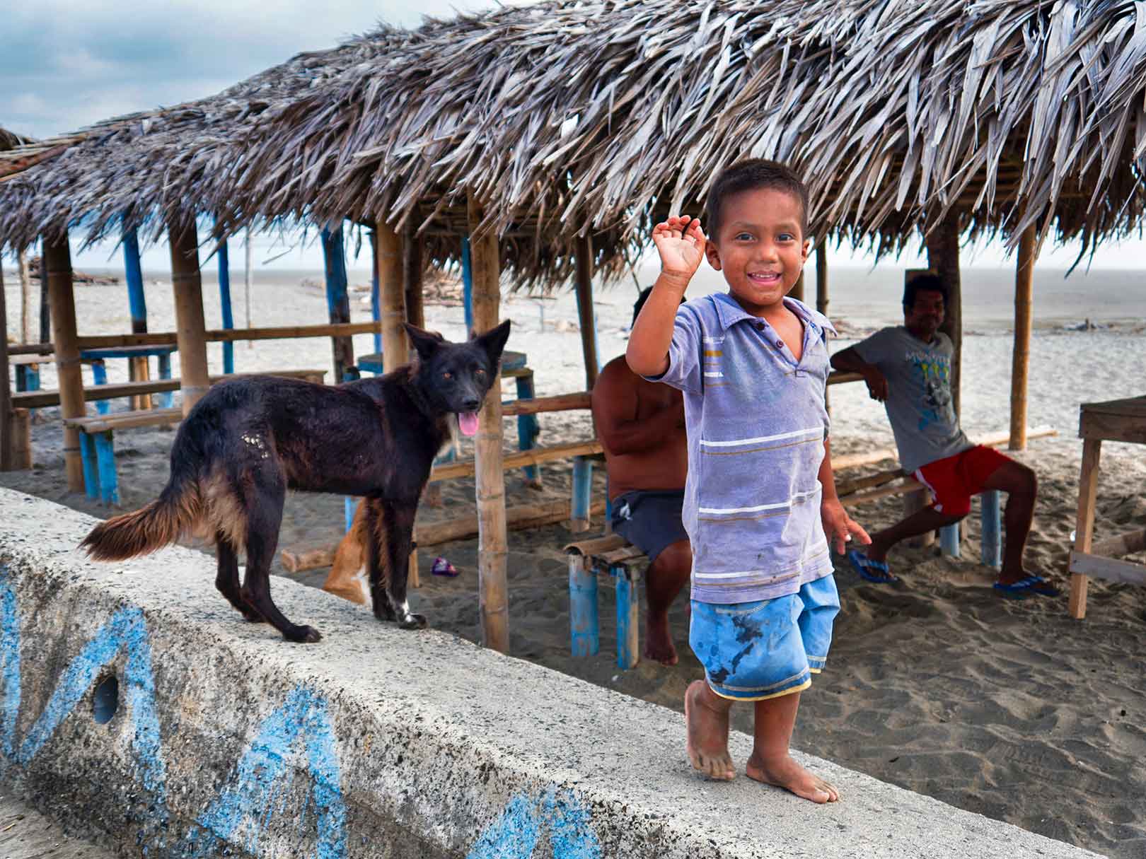 A small boy in blue shorts waving at the camera with his black dog standing on a concrete rail on a beach in Canoa, Ecuador.
