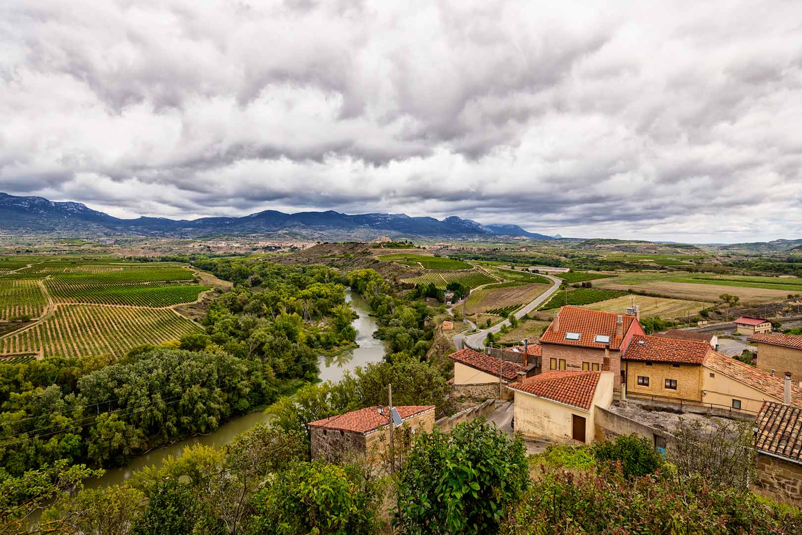 Aerial view of a rural landscape with a winding river, vineyards, scattered houses, and a cloudy sky.