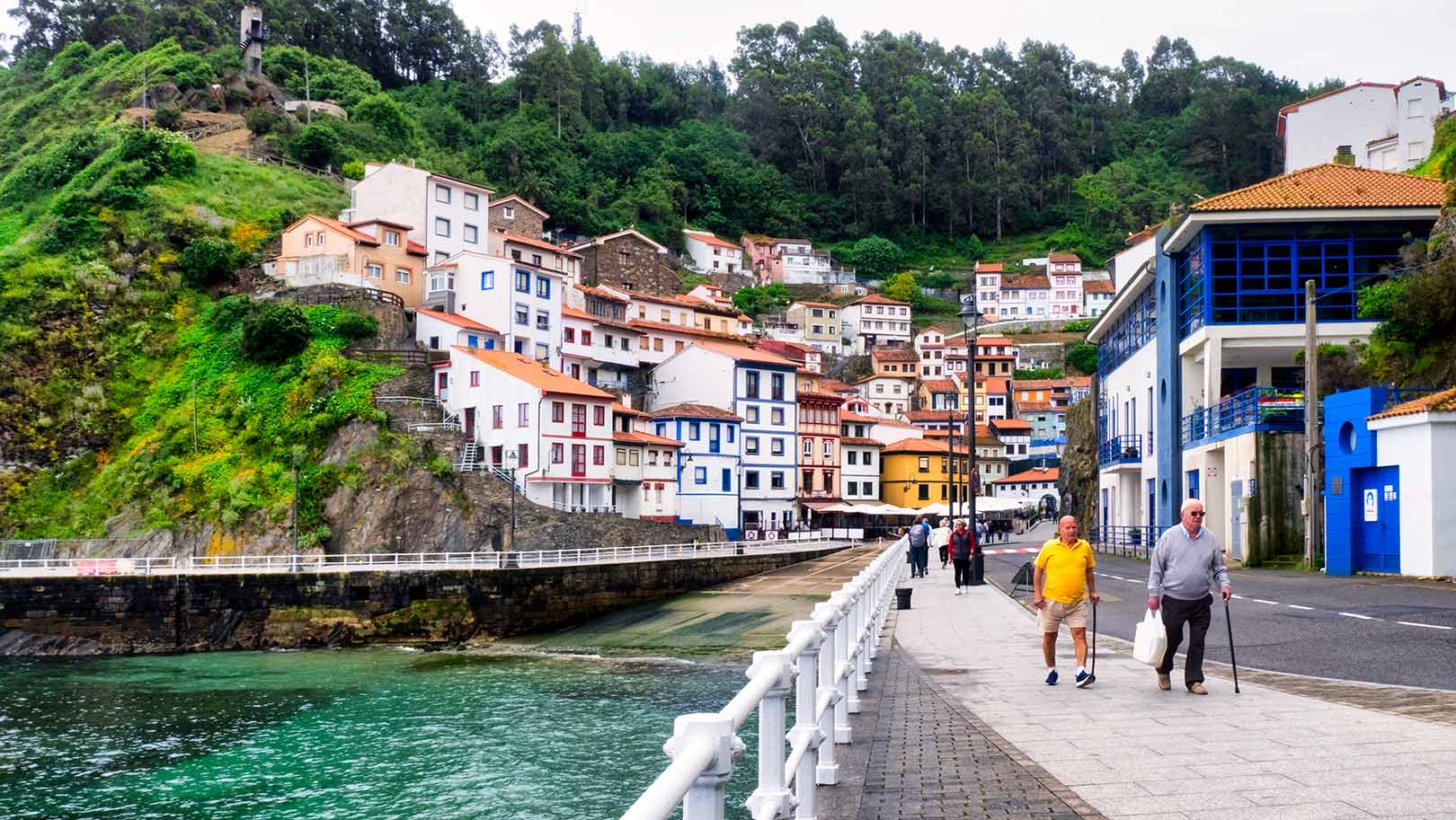 Two men walk along a concrete pier with canes in a small town with white washed buildings with colorful accents along a ridge in the distance.