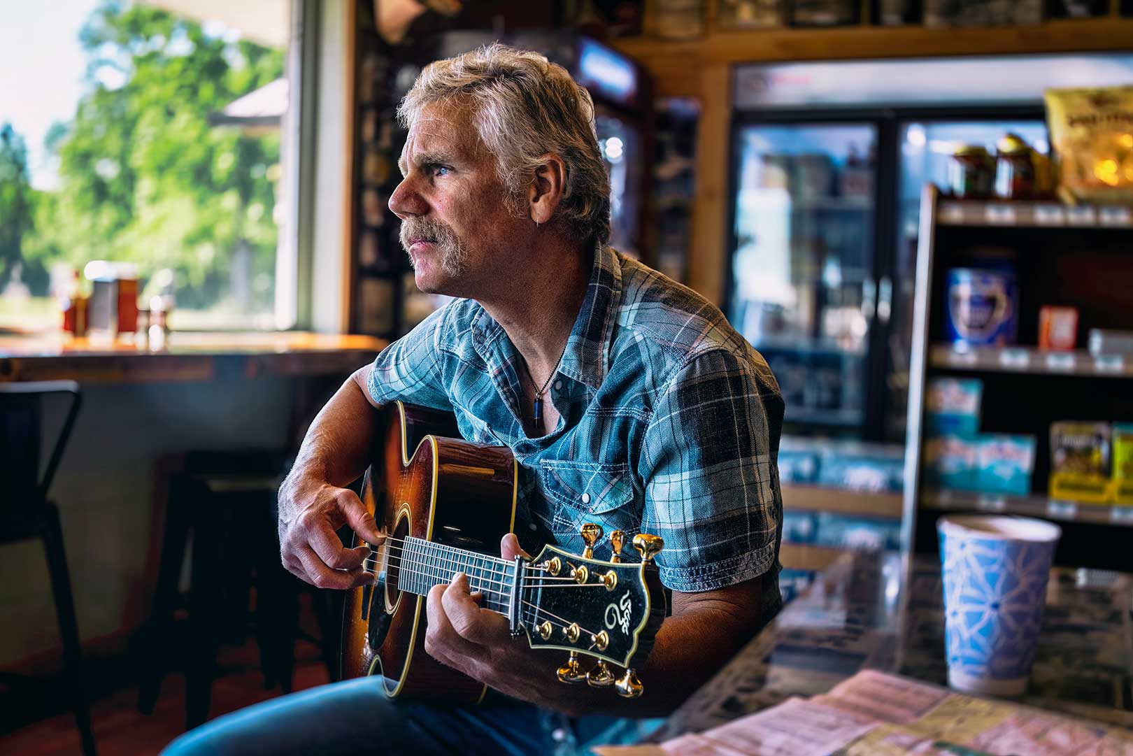 Man with gray hair and mustache playing an acoustic guitar in a casual indoor setting, wearing a plaid shirt. A table and items in the background.