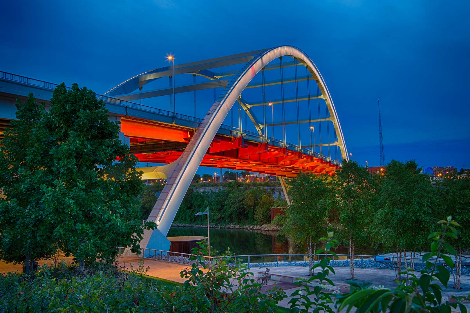Illuminated arch of the Nashville Korean War Veterans Memorial bridge with red accents spans over a river, surrounded by trees, under a twilight sky.