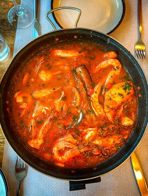A pot filled with seafood stew, including shrimp and mussels, in a rich tomato-based sauce, set on a table with a white plate and silverware.