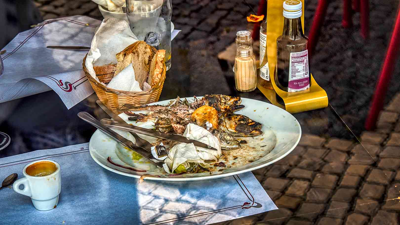 Empty fish plate with bones, cutlery, basket of bread, bottle of sauce, salt shaker, and an espresso cup on a table with cobblestone reflections.
