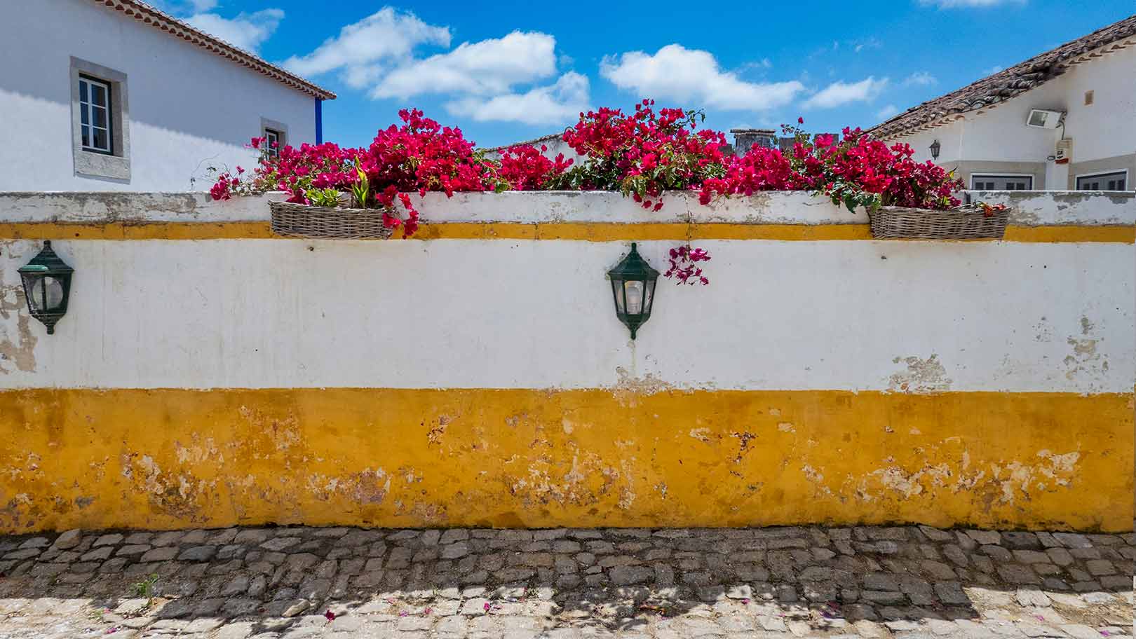 A stone pathway leads to a white and yellow wall with planters of red flowers on top. Two green lanterns are mounted on the wall. White buildings and a blue sky are in the background.