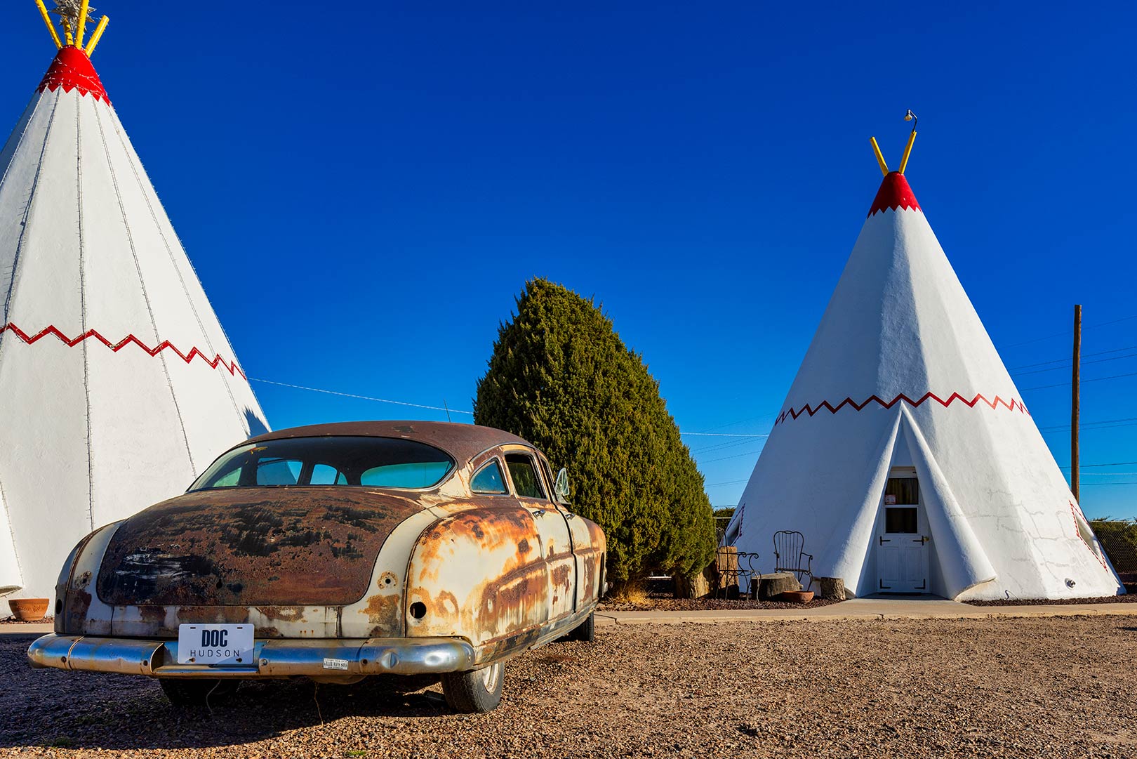 An old, rusty car is parked in front of two teepee-shaped motel rooms at the Wigwam Motel under a clear blue sky.
