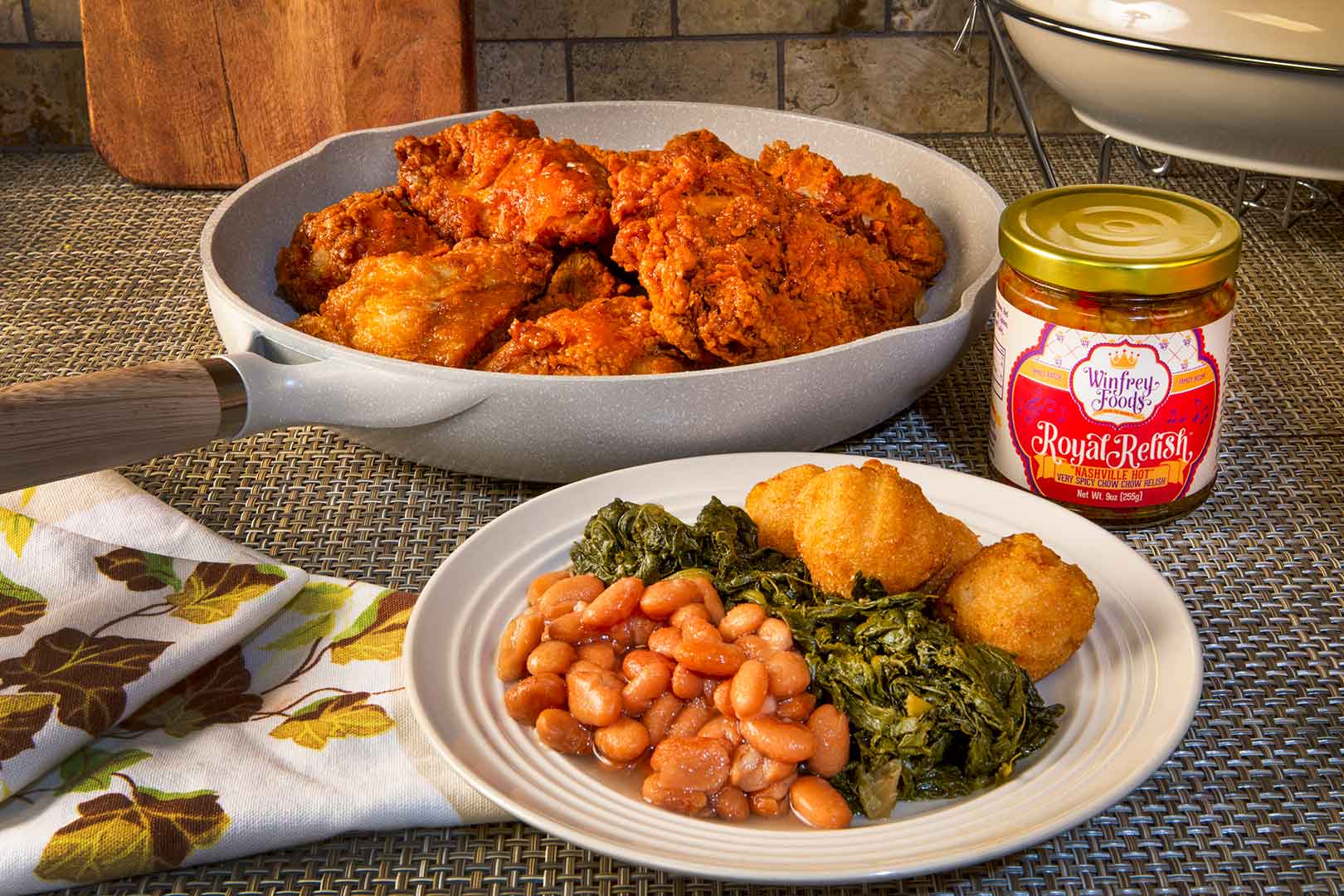 A plate with beans, greens, and hush puppies next to a pan of fried chicken and a jar of Royal Relish on a textured surface.