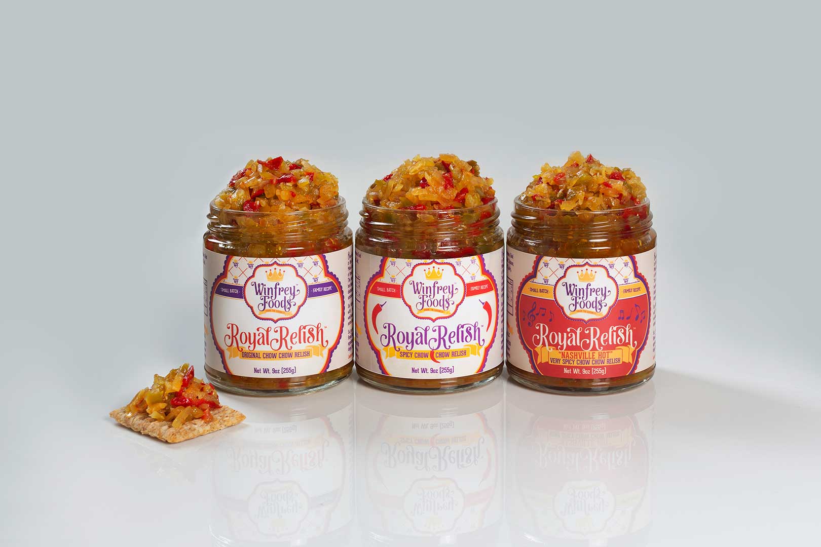Three jars of Royal Relish by Winfrey Foods in flavors, both, Red Pepper, and Classic Original, placed on a reflective surface with a relish-topped cracker beside them.